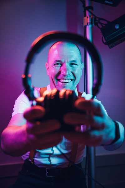 Ein lächelnder Mann im weißen Hemd und roter Fliege hält Kopfhörer in die Kamera, die in kräftiges lila und blaues Licht getaucht sind - und fängt die lebendige Energie von HochzeitsDj Christian für Franken und in Bamberg ein.