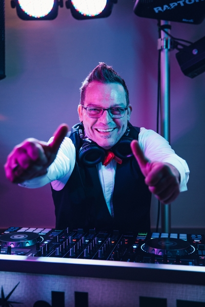 Ein lächelnder DJ mit Brille und Kopfhörern zeigt zwei Daumen nach oben hinter einem DJ-Mischpult, während im Hintergrund bunte Lichter leuchten - das ist HochzeitsDJ Ecky, der Energie und Spaß in jede Feier bringt.