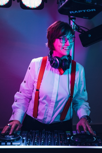 Die lächelnde Hochzeits-DeeJane Evi mit Brille, weißem Hemd mit roten Hosenträgern und Kopfhörern um den Hals steht an einem DJ-Mischpult in bunter rosa und blauer Beleuchtung.