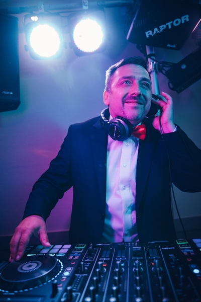HochzeitsDJ Marco für die Party in Franken und Thüringen steht lächelnd im Anzug und roter Fliege hinter dem DJ-Mixer, hält Kopfhörer ans Ohr und genießt die Stimmung von der Bühne.