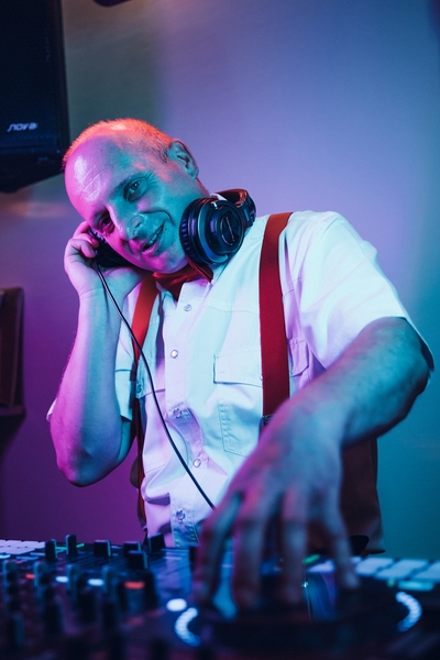 Ein lächelnder Mann mit weißem Hemd, roten Hosenträgern und Kopfhörern steht unter bunten Bühnenlichtern an einem DJ-Mischpult und scheint es zu genießen, als Hochzeits-DJ in Franken Musik aufzulegen.