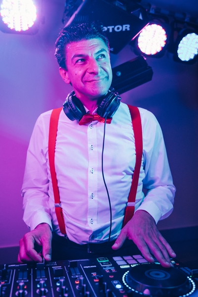 Ein lächelnder DJ für Hochzeit in Franken, gekleidet in ein weißes Hemd, rote Hosenträger und eine Fliege, steht an einem DJ-Controller mit Kopfhörern um den Hals, beleuchtet von bunten Bühnenlichtern.
