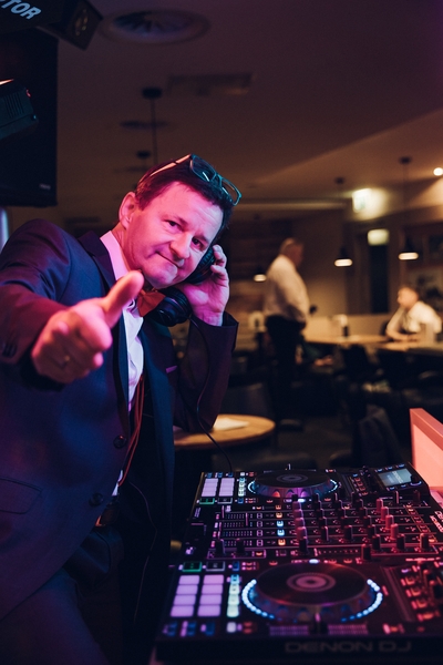 Ein DJ für Hochzeit in Thüringen, stilvoll im Anzug und mit Sonnenbrille, steht hinter einem DJ-Mischpult, hält Kopfhörer ans Ohr und zeigt in die Kamera in einer lebhaften, schwach beleuchteten Innenumgebung.
