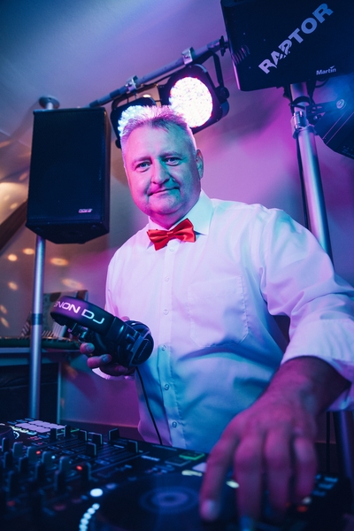 HochzeitsDJ Torsten mit weißem Hemd und orangefarbener Fliege steht an einem DJ-Pult und hält Kopfhörer in der Hand, während im Hintergrund bunte Bühnenlichter und DJ-Geräte zu sehen sind.